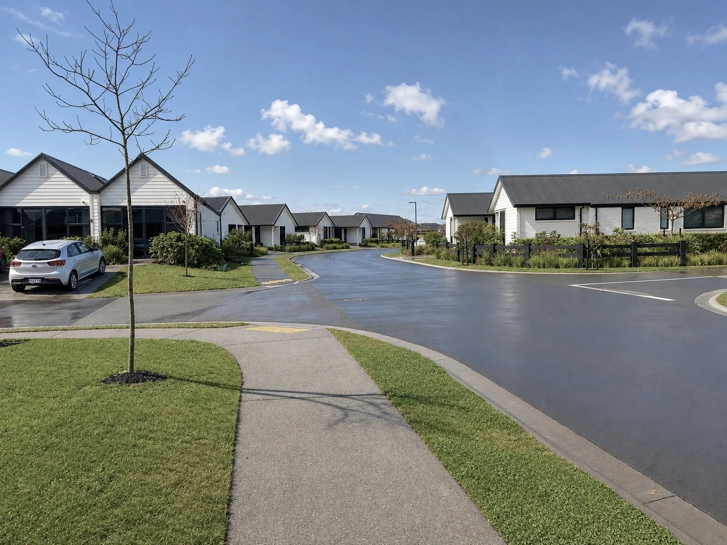 A suburban neighborhood street with modern white houses, black roofs, and well-maintained lawns. The street is wet, possibly from recent rain, with a clear blue sky and some clouds. There are a few leafless trees, cars parked along the driveway, and a sidewalk running parallel to the street.