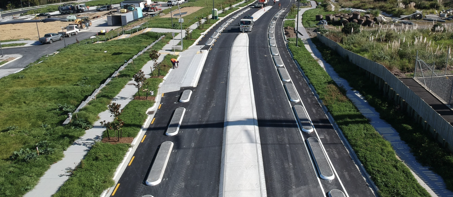 A new paved road is under construction with a worker and a small truck on it. On the left side, there are green grassy areas and newly planted small trees. On the right side, there is a sidewalk with shrubs and a fencing area. In the background, there are parked cars and construction materials.
