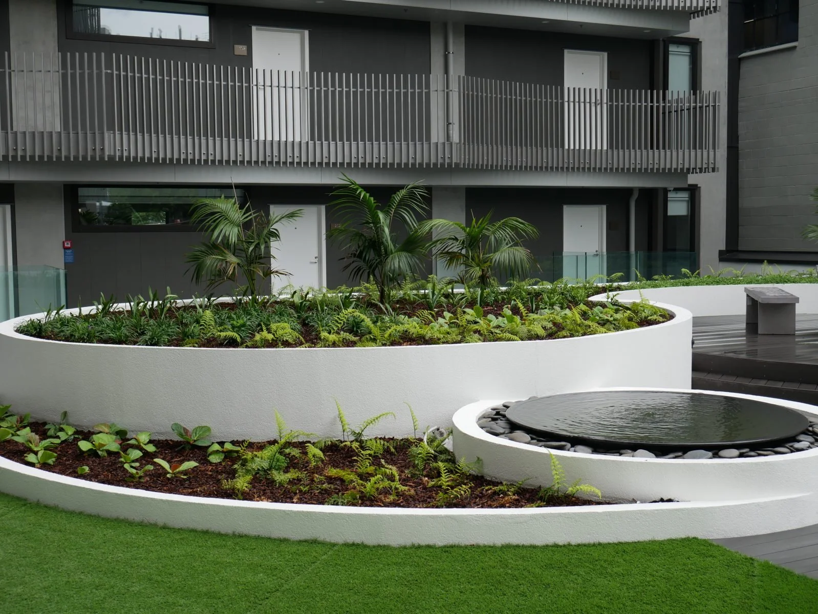 Modern urban courtyard with lush green plants, a white curved planter, and a water feature, adjacent to a contemporary building with gray exterior and balconies.