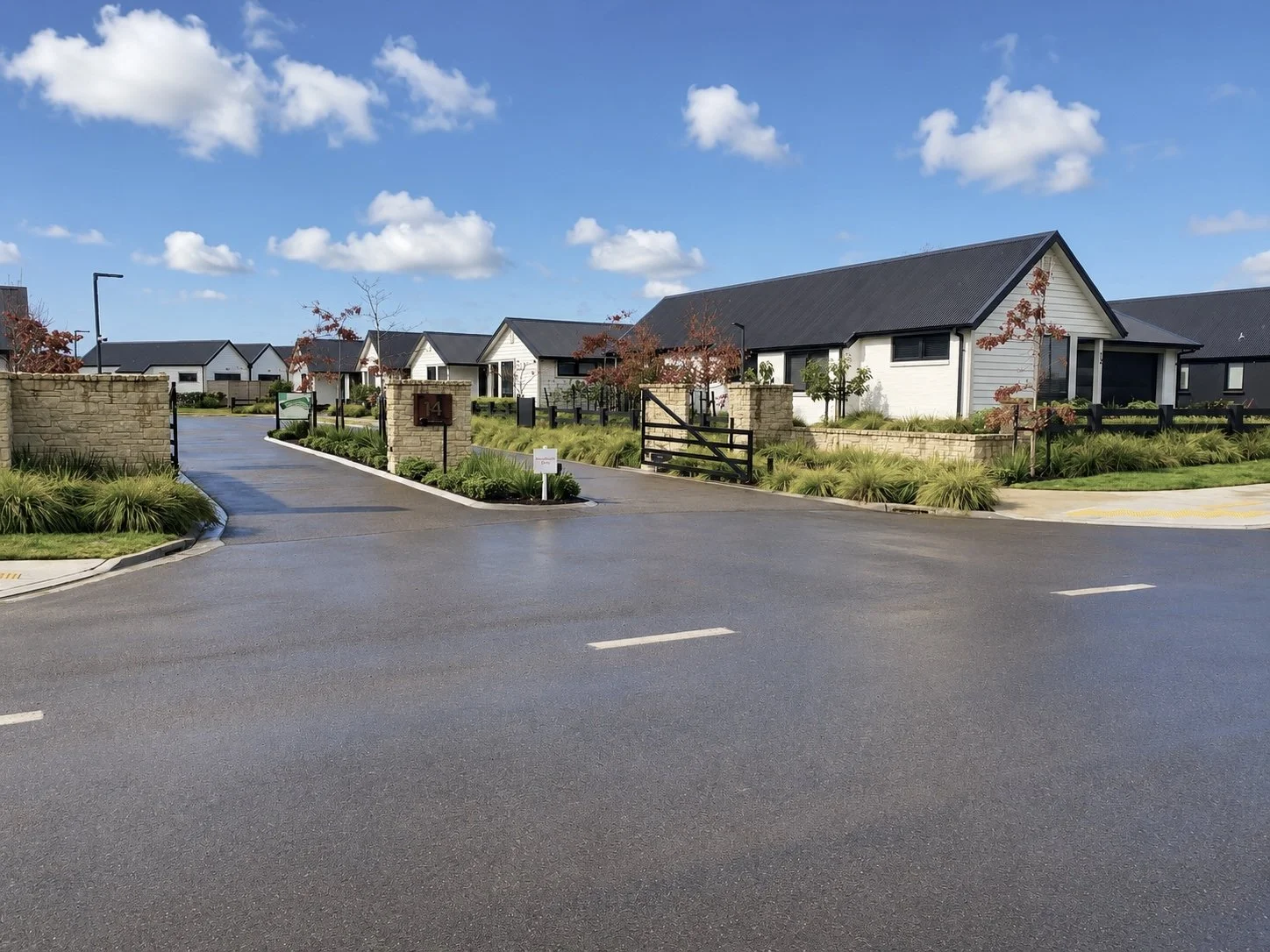 A residential gated community with modern white and black houses, landscaped yards, and a paved entrance road under a blue sky with clouds.