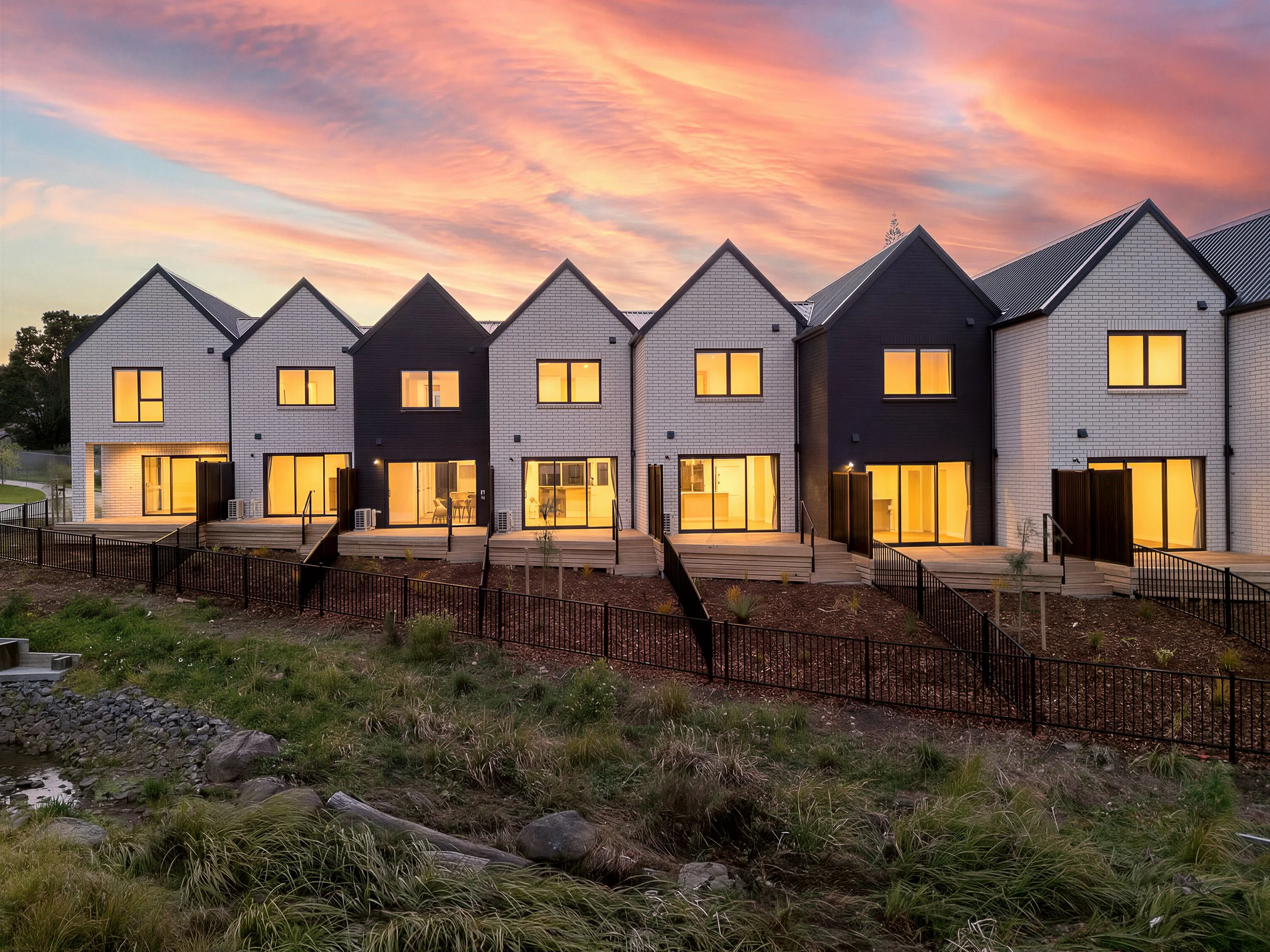 Modern row of townhouses with white and black exteriors, large illuminated windows, and wooden deck patios, set against a sunset sky with pink and orange clouds.
