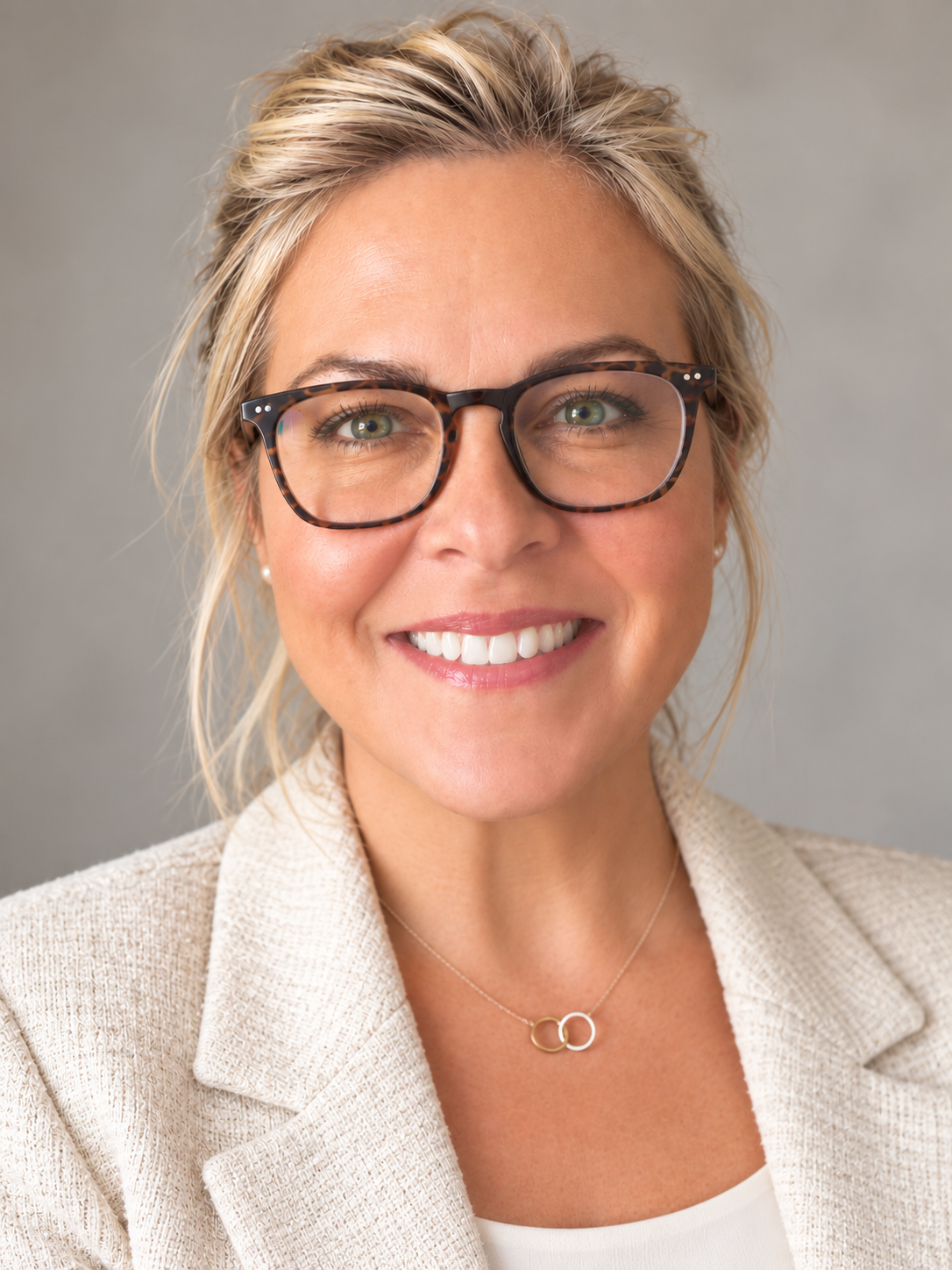Close-up of a woman with blonde hair, glasses, and a smile, wearing a beige blazer and necklace, against a neutral background.