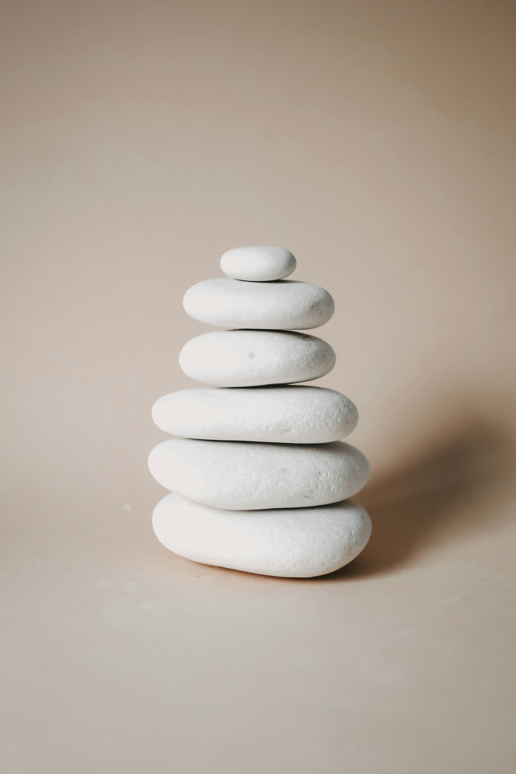 Stack of seven smooth, white stones arranged in descending size order against a neutral background with a soft reflection on a surface.
