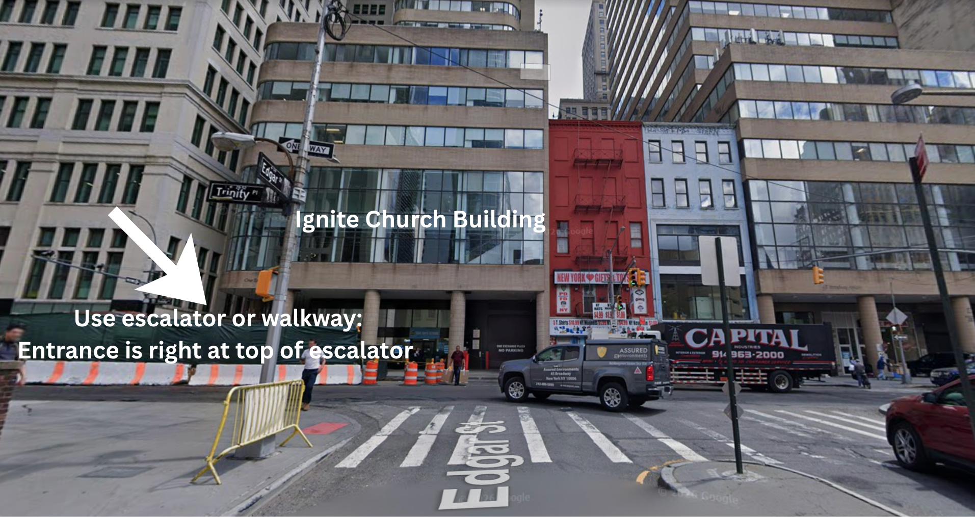 A city street scene showing a large modern office building labeled Ignite Church Building and a red building with a fire escape. There are parked trucks and cars on the street, pedestrian crosswalks, and street signs including one for Trinity Place. An arrow points to an entrance of the Ignite Church Building indicating the use of an escalator or walkway, with signage noting the entrance is at the top of the escalator.