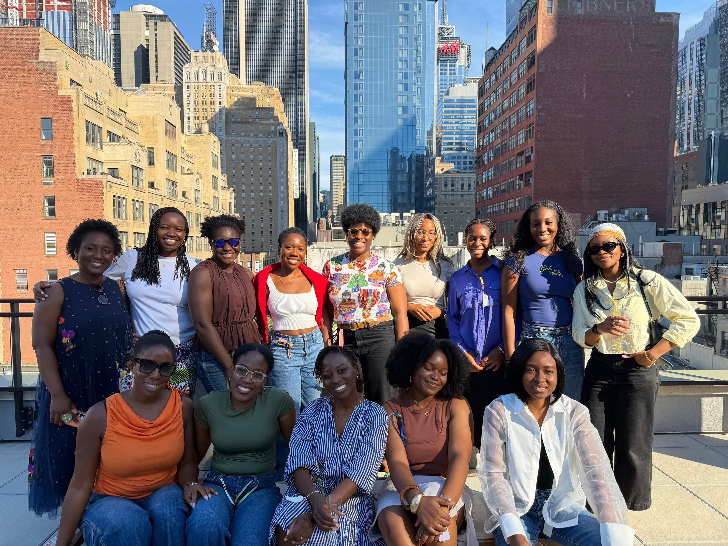 A group of women standing together outdoors in front of a city skyline with tall buildings and blue sky, smiling at the camera.