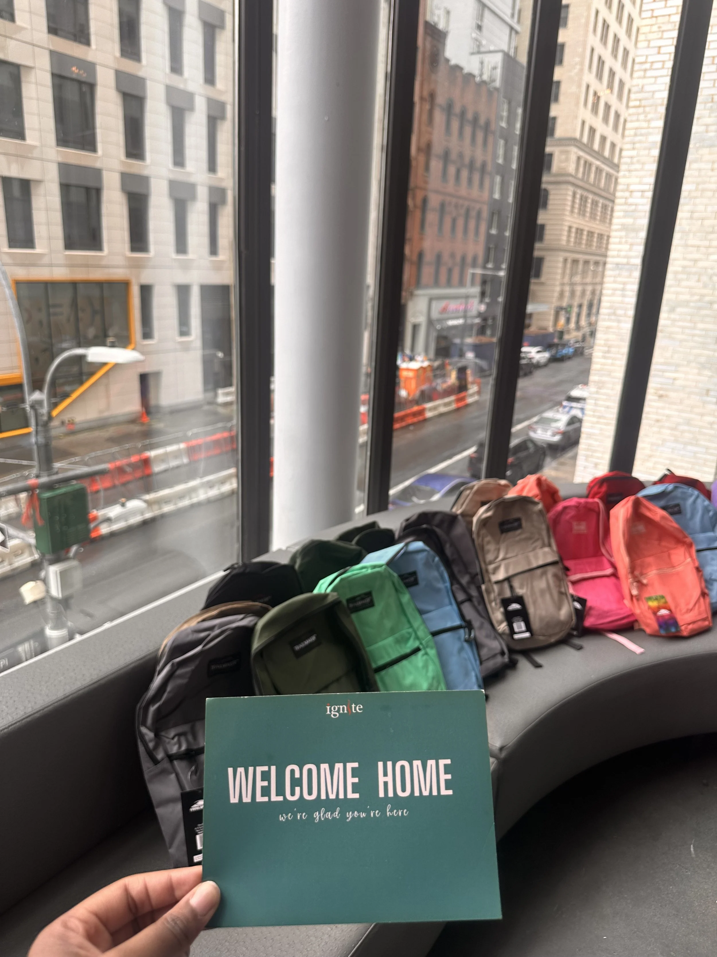 A card that says "Welcome Home" with a row of backpacks in the background.