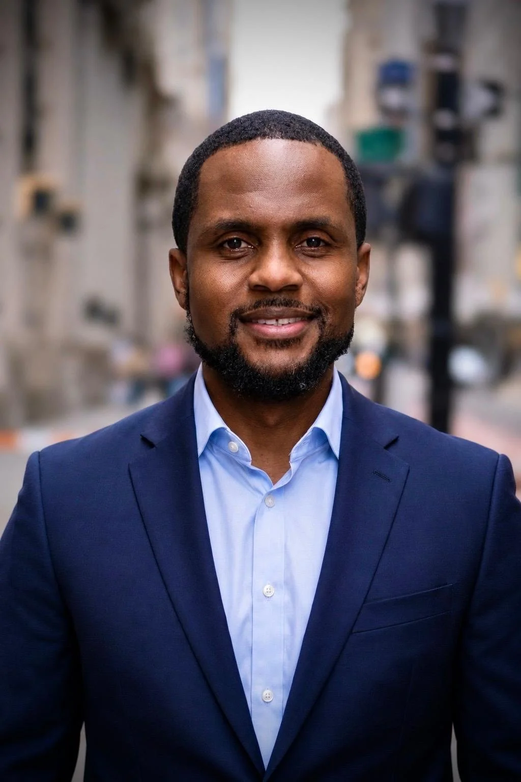 A man in a navy suit and light blue shirt smiling outdoors in an urban area.