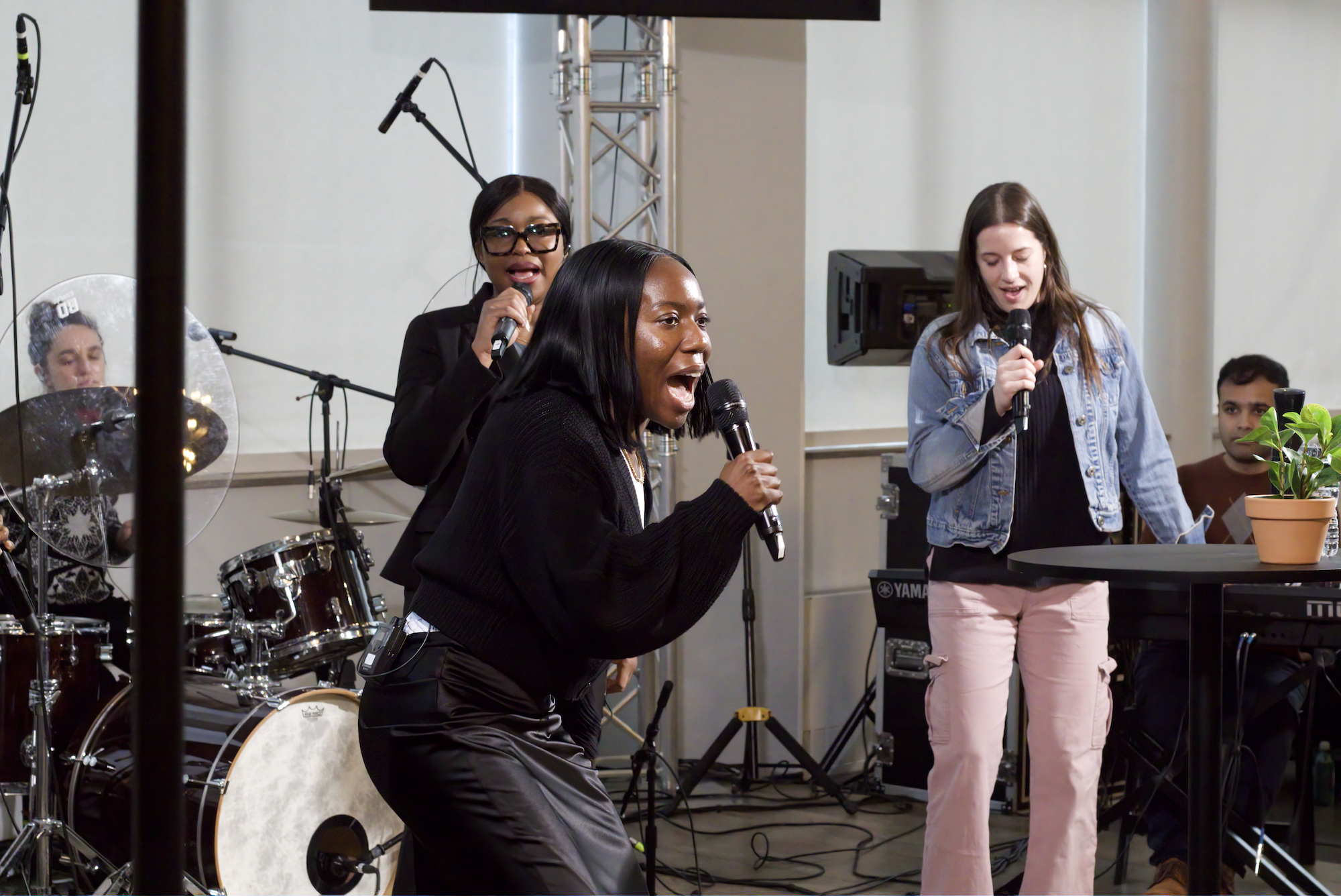 Worship team members performing live, including a woman singing into a microphone, another woman singing with a microphone, and a drummer playing drums, with musical instruments and equipment in the background.