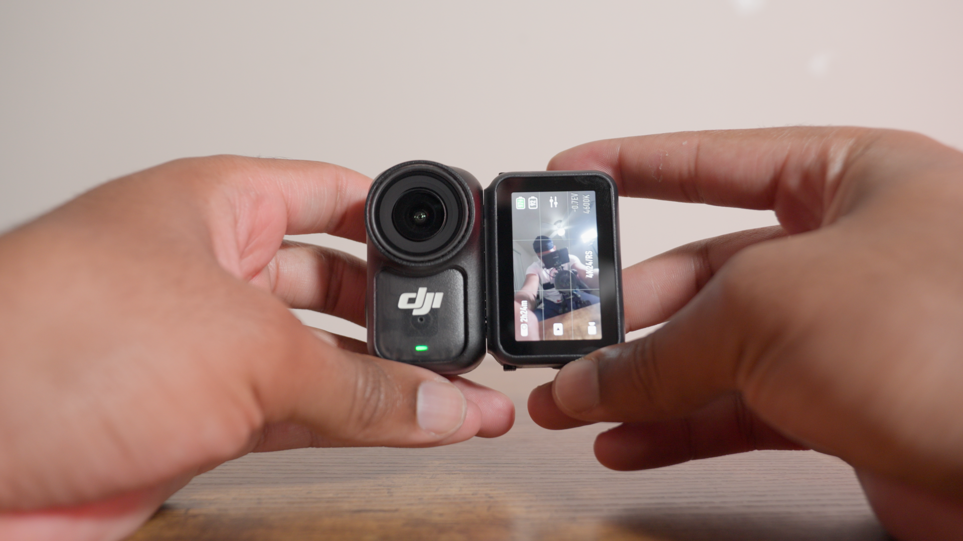 A person holding a black DJI action camera with a small built-in display screen showing a reflection of the person taking the photo on a wooden table against a plain background.