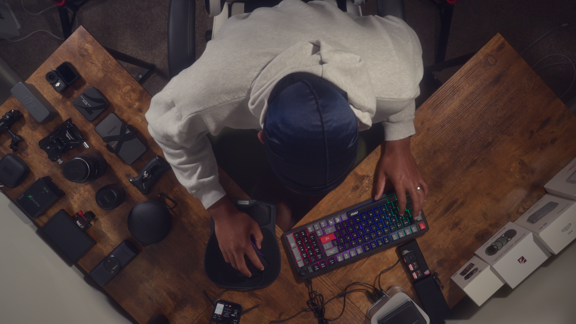 Top-down view of a person working at a wooden table with electronic devices, cameras, and accessories, typing on a colorful keyboard.