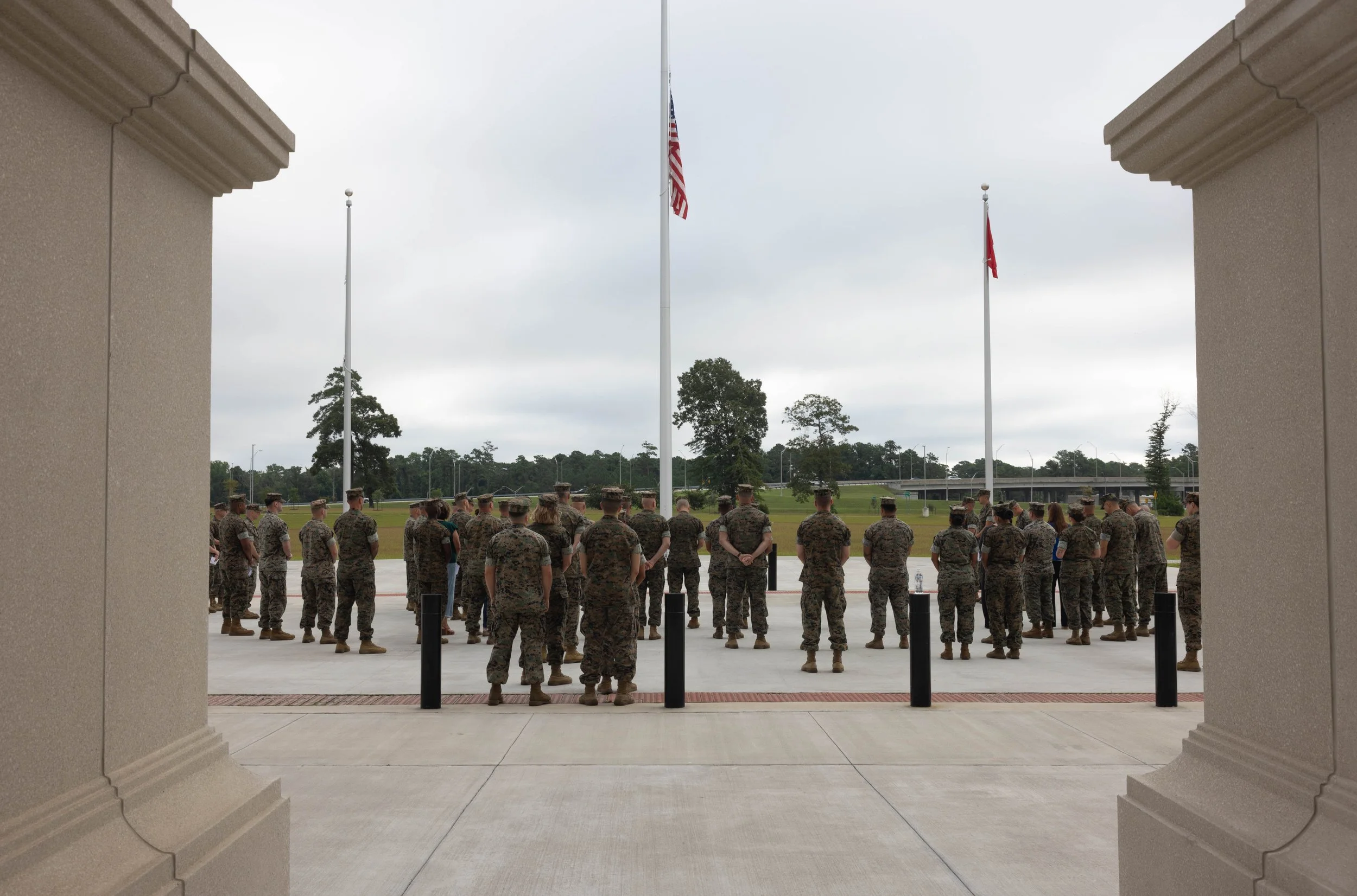 A group of soldiers standing in formation outside in a military ceremony, viewed through an architectural opening with two beige stone columns on either side and two flagpoles with American and other flags in the background under a cloudy sky.