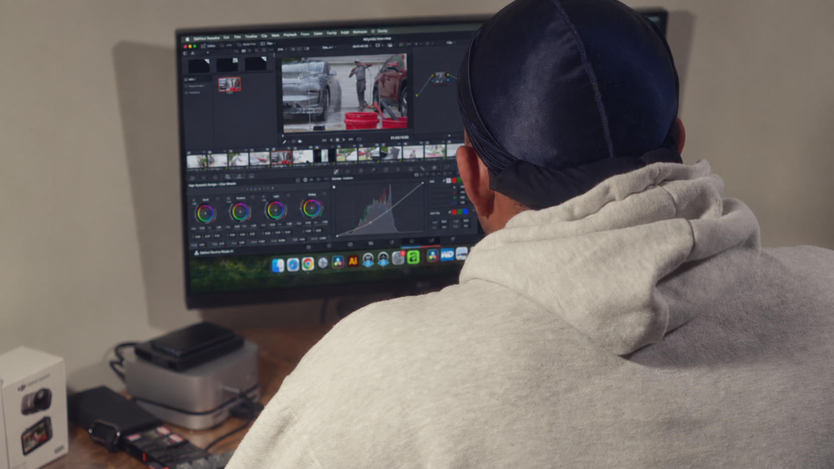 A person wearing a hoodie and cap working on video editing on a large monitor, displaying various editing tools and footage. The workspace includes a desk with electronic devices and accessories.