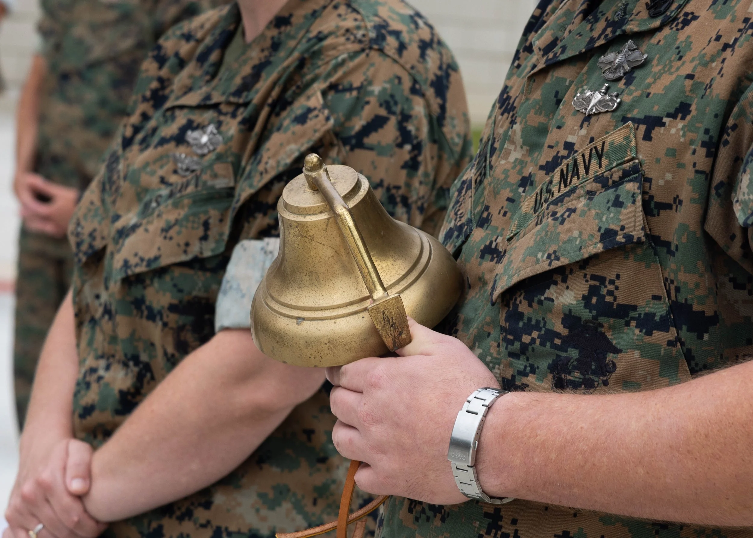 Close-up of a person in U.S. Navy uniform holding a brass bell, with other individuals in similar uniforms visible in the background.