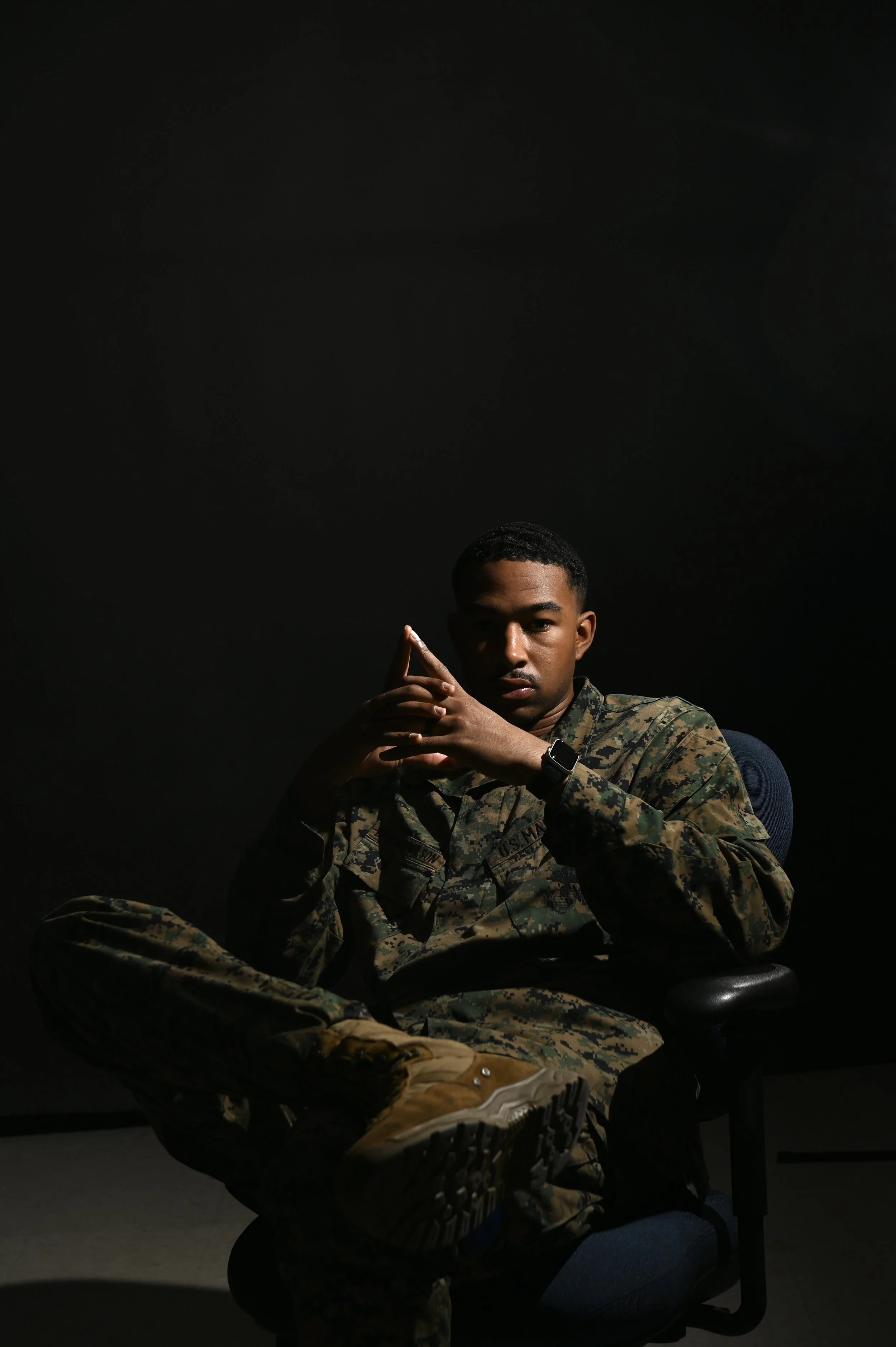 A young man wearing a military camouflage uniform sitting on a blue office chair against a dark background, with his hands clasped in front of him and wearing a wristwatch.