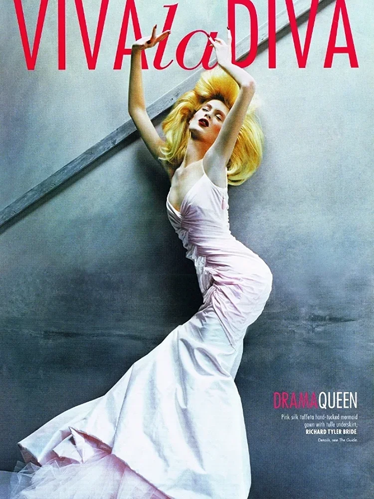 A woman with voluminous blonde hair in a white gown with a pink silk taffeta mermaid style skirt, posing with arms raised and head tilted back against a gray wall with large red text reading 'VIVALA DIVA' at the top and smaller pink and white text at