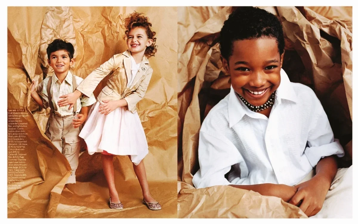 Two children, a boy and a girl, standing and smiling against a brown paper background, with another boy sitting inside crumpled brown paper, smiling and looking at the camera.