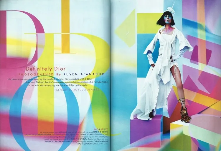 Model wearing a white dress with high slit, decorated with lace-up shoes, standing on a colorful geometric background with pastel shades of blue, pink, yellow, and purple.