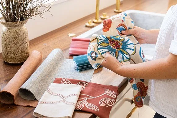 Person holding a piece of fabric with a floral pattern beside various fabric rolls and sewing tools on a wooden table.