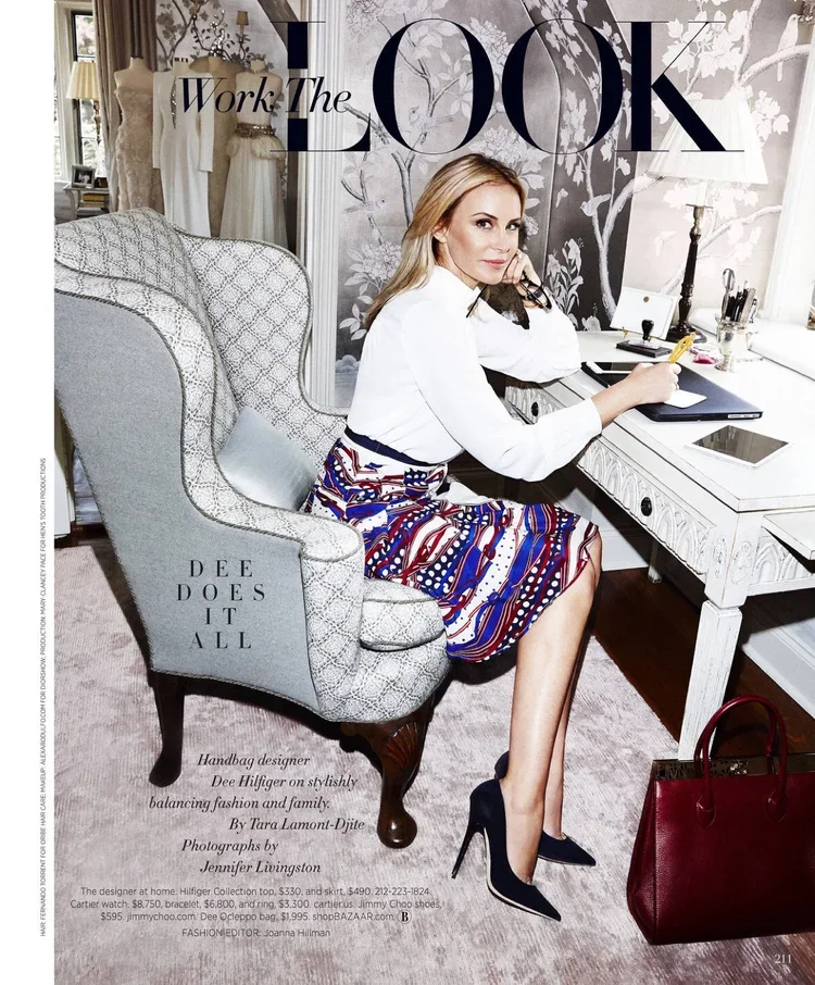 A woman sitting on a stylish armchair at a white desk, holding a pen, with a laptop, tablet, and various office supplies. She is dressed fashionably with a white top and a colorful patterned skirt, wearing high heels. The background shows a wall with