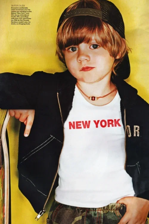A young boy with shoulder-length brown hair is wearing a black trucker hat backwards, a white T-shirt with 'NEW YORK' written in red, a black zip-up jacket, and camouflage pants. He is pointing at his shirt with his right thumb and standing against a