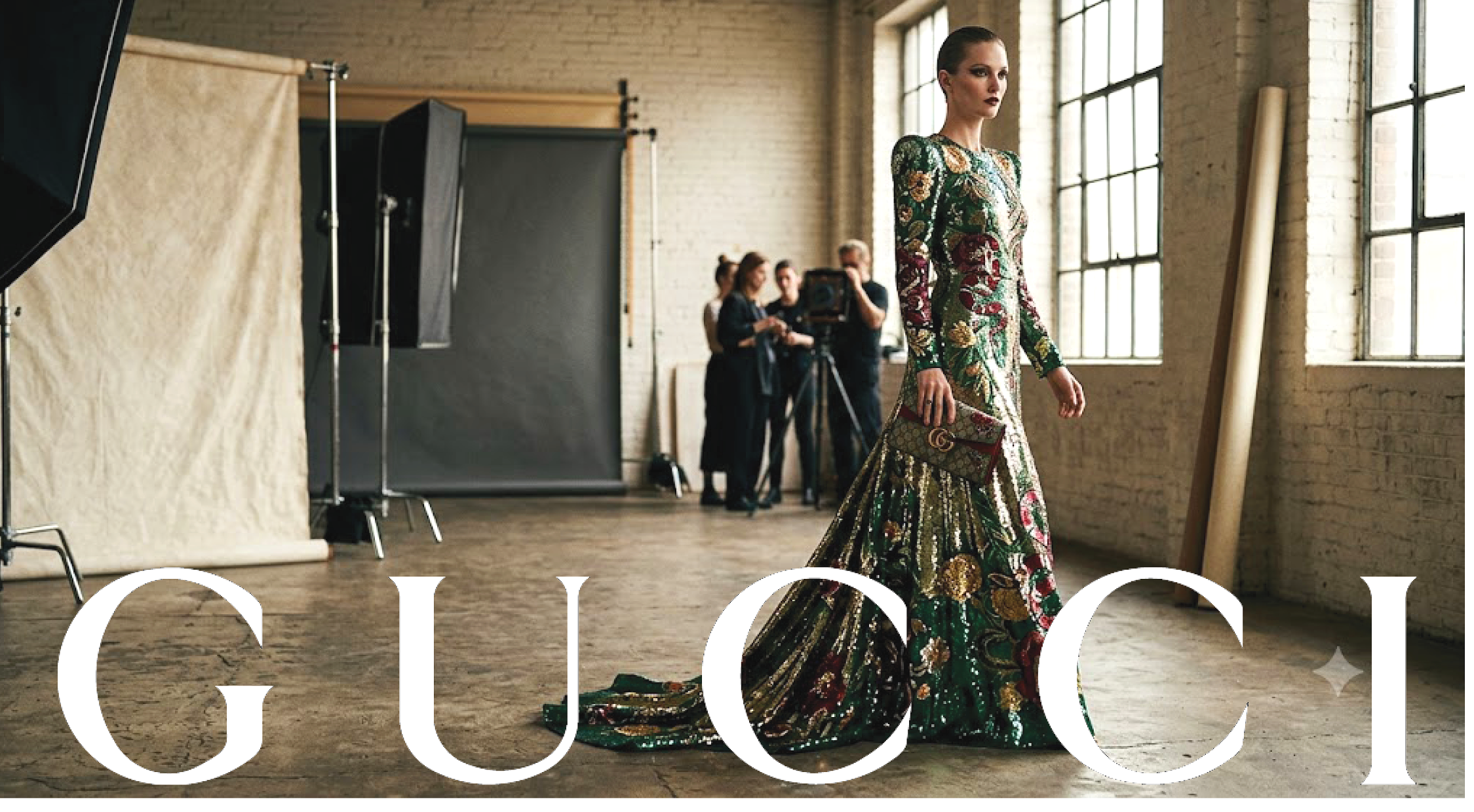 A female model wearing an ornate, floral-patterned gown with long sleeves, holding a matching clutch purse, in a photography studio with large industrial windows and a beige backdrop.