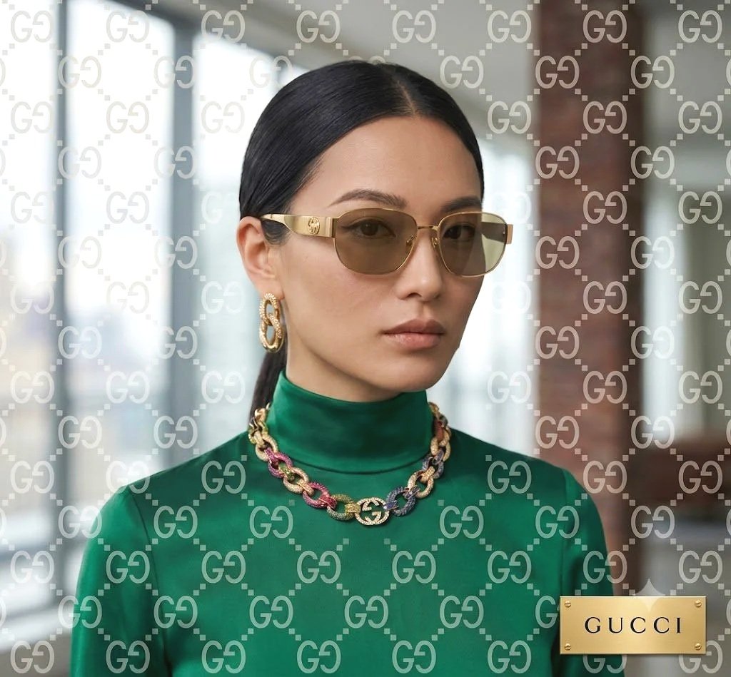 A woman wearing gold sunglasses, gold earrings, a multicolored chain necklace, and a shiny green turtleneck top standing in front of a glass window with a reflection. The photo has a Gucci branding overlay.