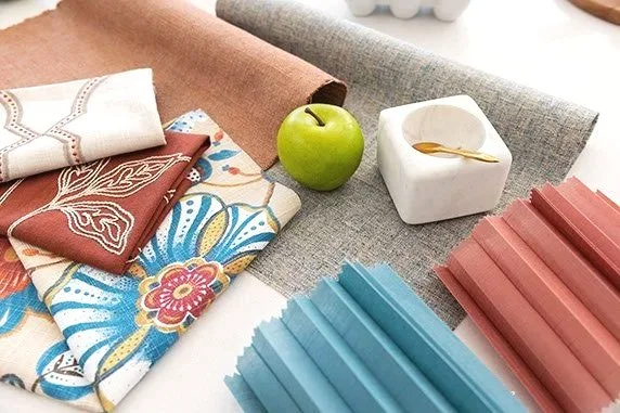Fabrics with floral, paisley, and solid patterns, a green apple, a small white dish, and colored paper or fabric strips on a gray surface.