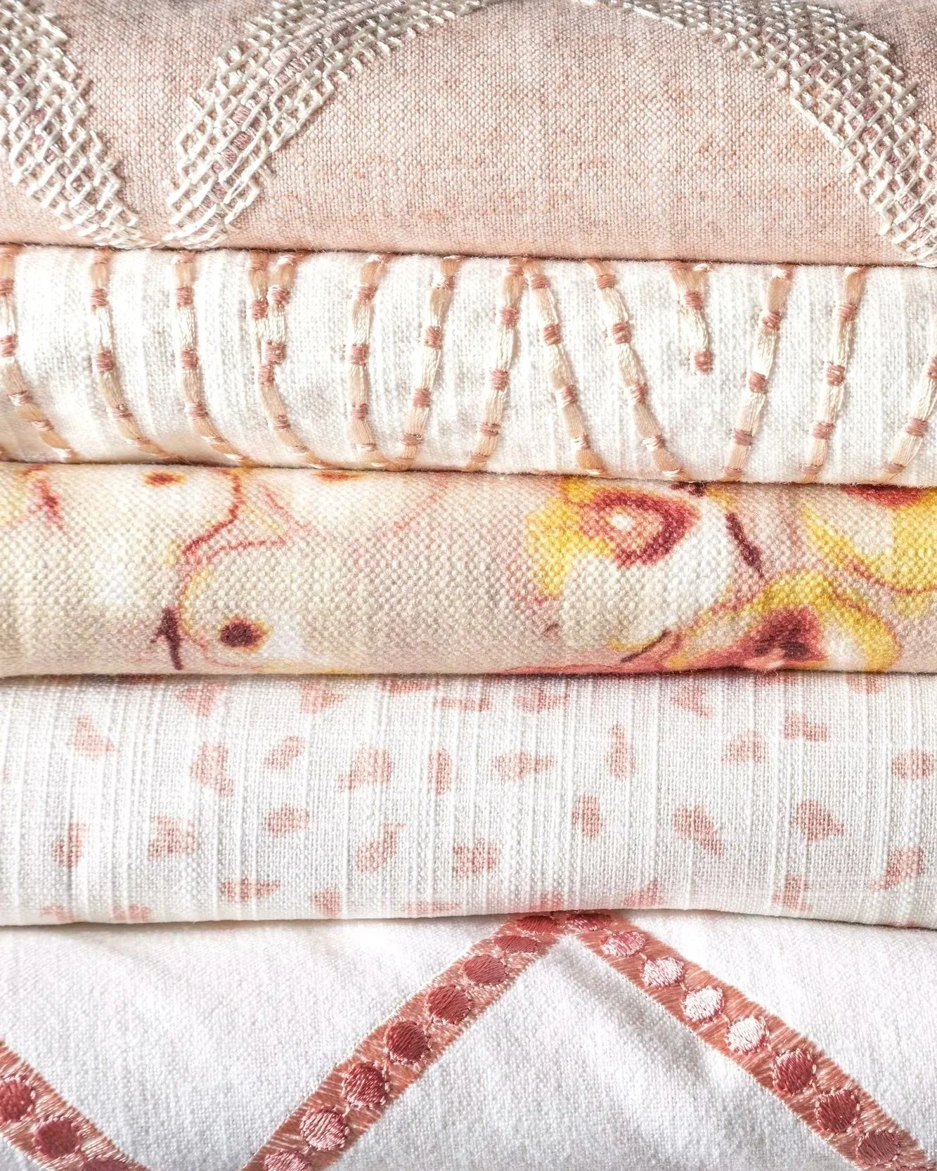 A stack of four folded fabrics with pink and peach tones, featuring various embroidered patterns including lattice, floral, and geometric designs.
