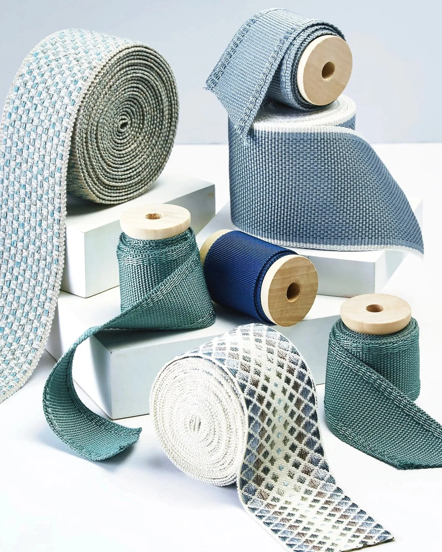 Various rolls of woven fabric in different shades of blue, gray, and white, displayed on white blocks and surfaces.
