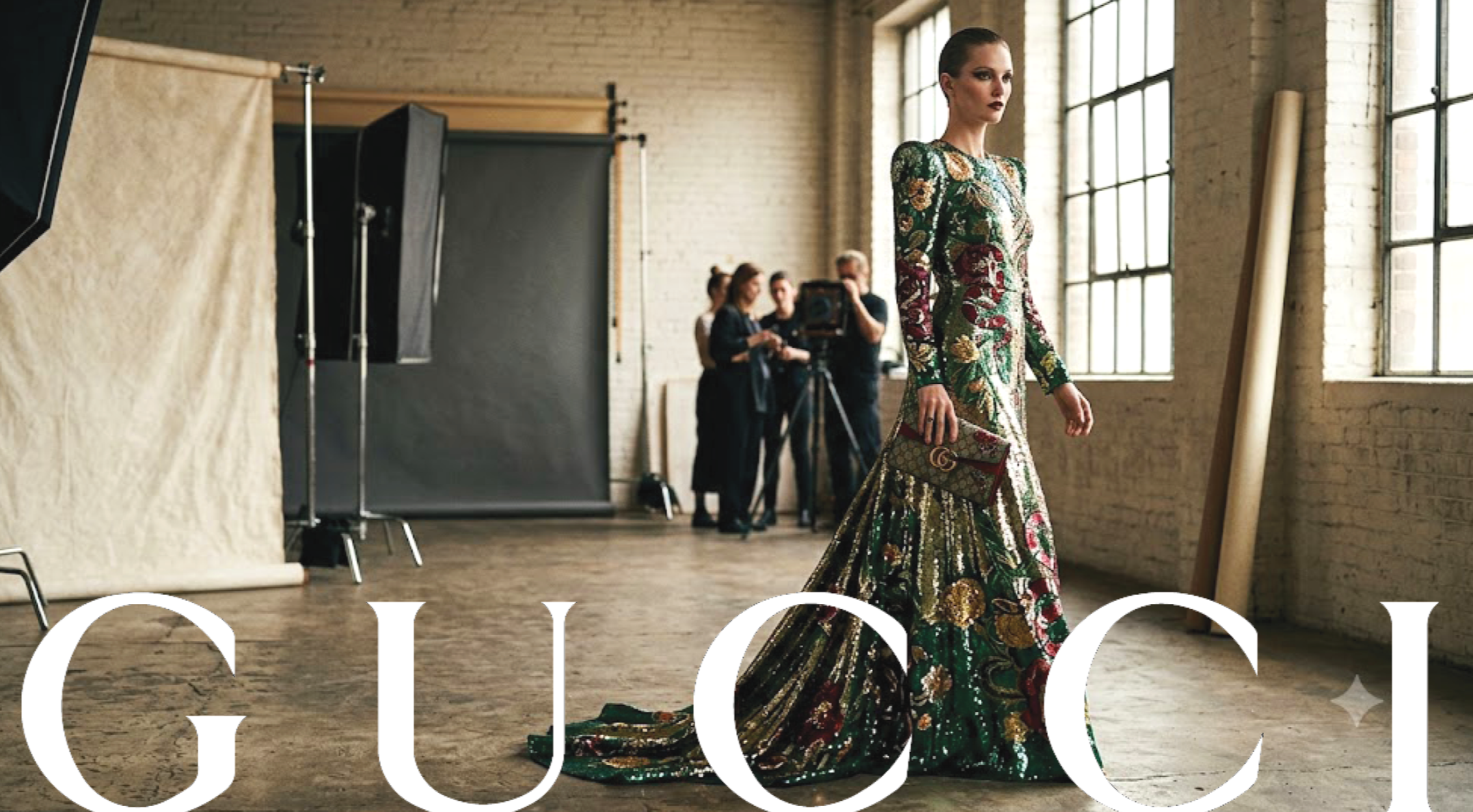 A female model wearing an ornate, floral-patterned gown with long sleeves, holding a matching clutch purse, in a photography studio with large industrial windows and a beige backdrop.