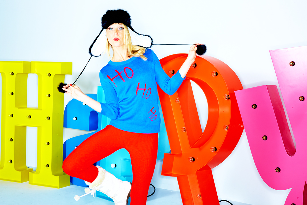 Woman wearing a blue sweater with 'Ho Ho Ho' written on it, red pants, white boots, and a black furry hat with ear flaps, standing in front of large colorful letter signs.