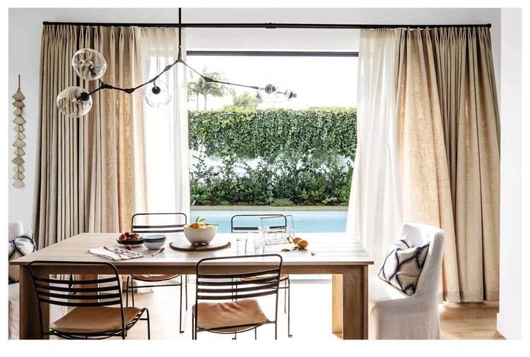 Dining room with a wooden table, four chairs, and a white armchair, large window with cream curtains, greenery and pool visible outside, modern light fixture hanging from ceiling.