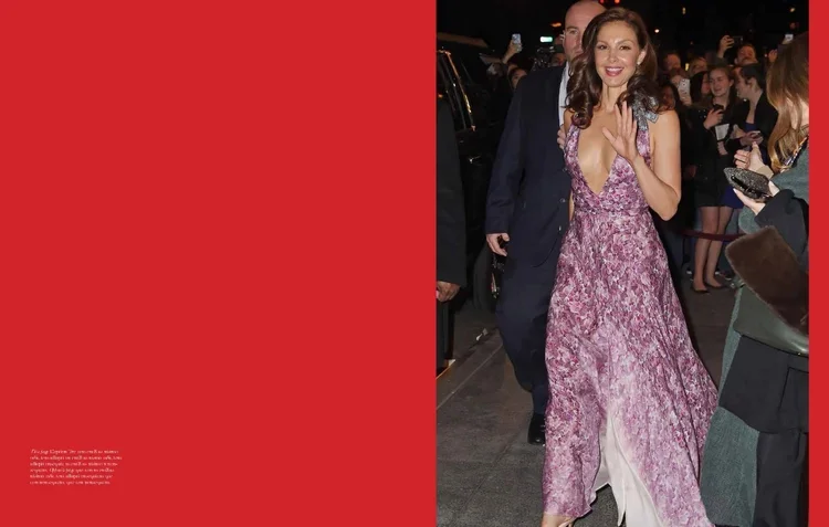 A woman in a pink lace gown walking on a city street at night, smiling and waving at the camera, surrounded by people and holding a small clutch.