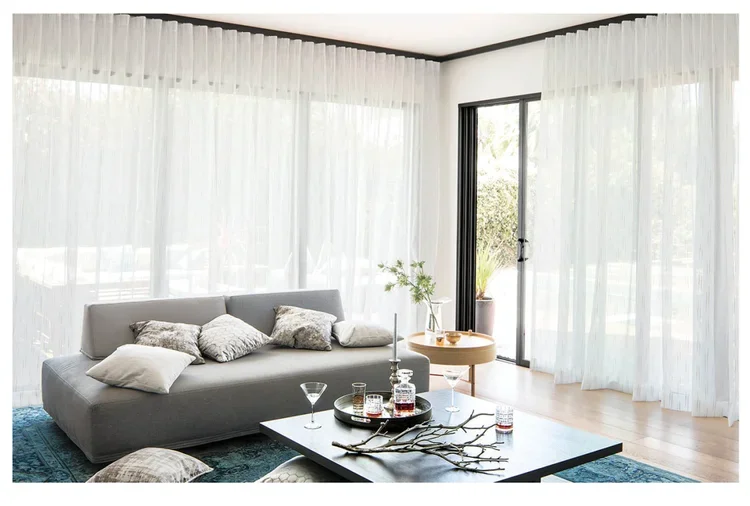 Modern living room with large windows covered by white sheer curtains, gray sofa with pillows, glass coffee table with drinks and decorative branches, round side table with a plant, and light wooden flooring.