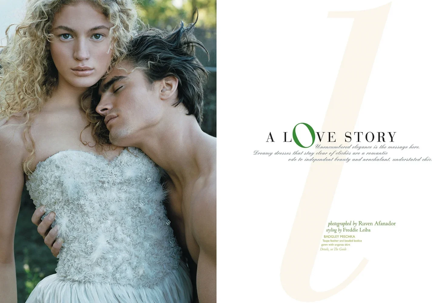 A romantic photograph of a woman with curly blonde hair and blue eyes, wearing a white textured dress, holding a shirtless man with dark hair, resting his head on her chest with his eyes closed, outdoors with blurred green foliage in the background. 