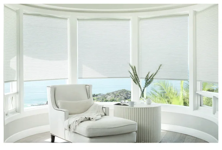 A cozy white armchair with a blanket draped over it next to a small round table with a vase of flowers and a cup, in a bright room with large bay windows showing an ocean view.