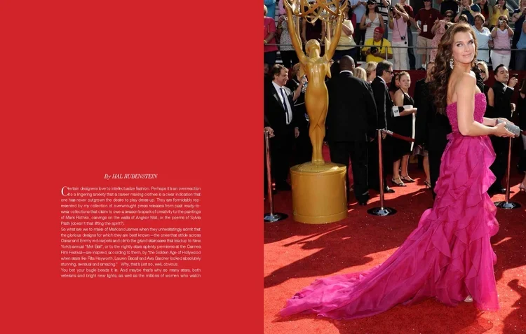 A woman in a pink strapless gown with a long train posing on a red carpet at an award event, with people and a large gold award statue in the background.