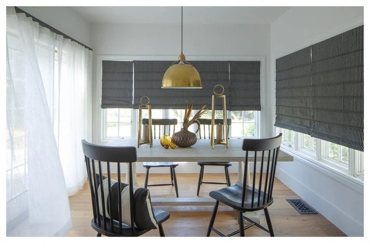 A dining area with a white table, four black chairs, and a decorative centerpiece with a vase and dried branches. Large windows with gray roman blinds and sheer curtains allow natural light into the room. A hanging gold pendant lamp is above the tabl