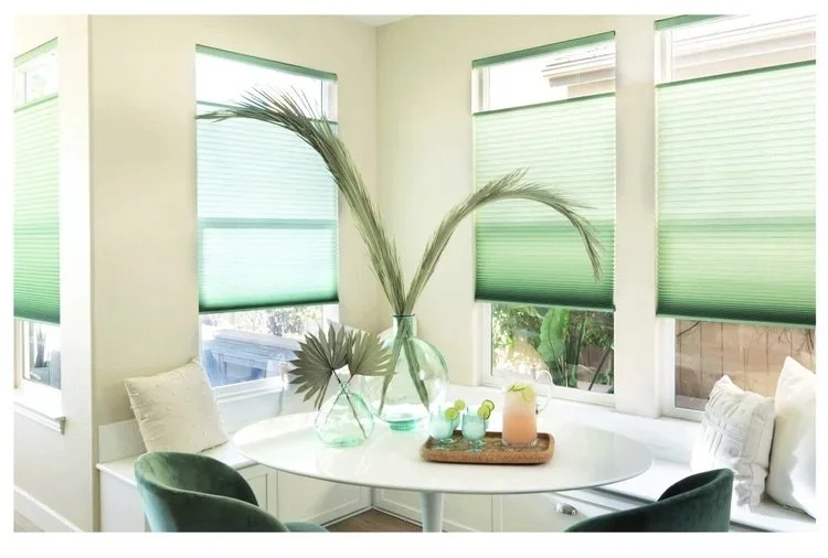 Sunlit dining nook with four windows covered by green blinds, decorated with a white oval table, two green chairs, and a glass vase with tall grass pampas on a tray with drinks and glassware.