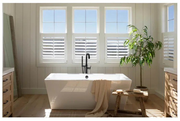 A bathroom with a large white bathtub in front of three windows with plantation shutters, a tall green plant to the right, and a small wooden table with soap in front of it.