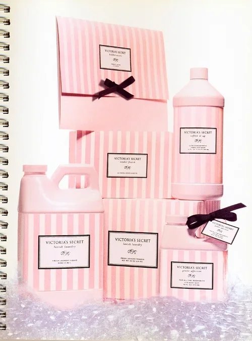 A collection of Victoria's Secret Pink laundry products, including a large pink bottle, a pink jug, and smaller containers, all with pink and white striped packaging and black bows, arranged with gift boxes.