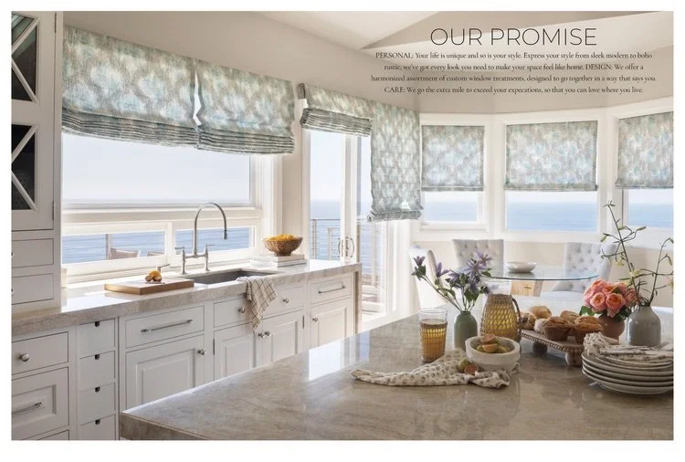 Bright coastal kitchen with white cabinets, granite countertops, and large windows with blue patterned Roman shades overlooking the ocean, decorated with flowers, plates, and baked goods on the island.