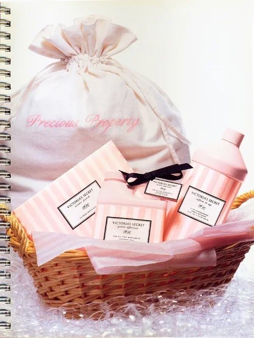 A gift basket with Victoria's Secret items including lotion, perfume, and a small box, wrapped in pink and white striped paper, with a cloth bag labeled 'Precious Property' in the background.