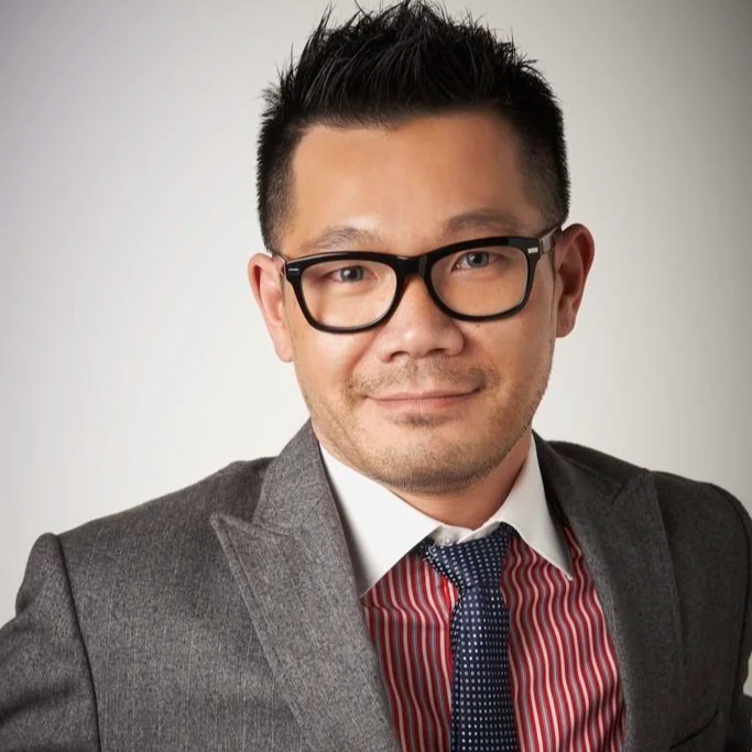 A man wearing black glasses, a grey suit, a white shirt, and a red and white striped tie, smiling at the camera against a plain background.
