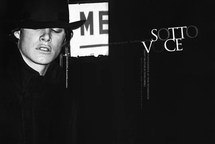 A black and white photograph of a woman wearing a black hat and coat, with eyes closed, standing in front of a dark background with white text reading 'SOTTO VOCE'.