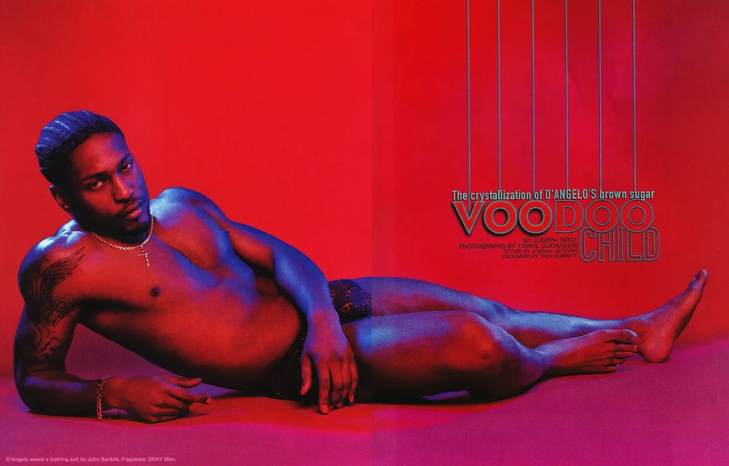D'Angelo reclined on a red background, wearing a bathing suit, with a gold chain and cross necklace, showcasing tattoos on his arm and body.