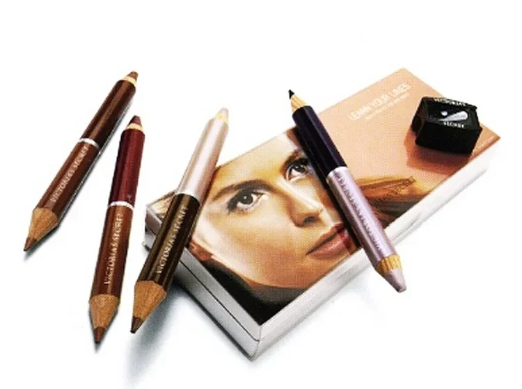 Makeup pencils, a makeup palette, and a face mirror with a woman's face reflected, arranged around a compact mirror or makeup case.