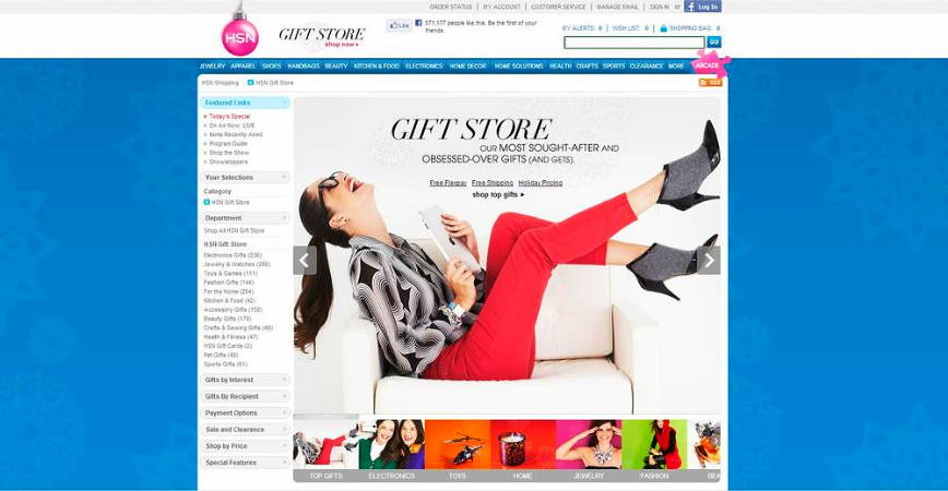 Screenshot of the HSN Gift Store website featuring a woman in red pants and black high heels sitting on a white sofa, laughing and holding a gift box. The website header includes navigation links and a shopping cart icon.