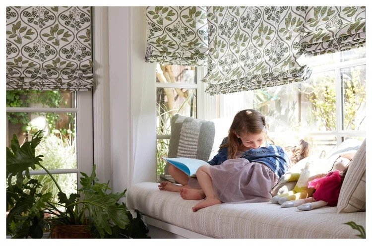 A young girl sitting on a window seat reading a book with her hands behind her head, surrounded by pillows and stuffed animals, in a bright room with patterned curtains and sunlight streaming in.