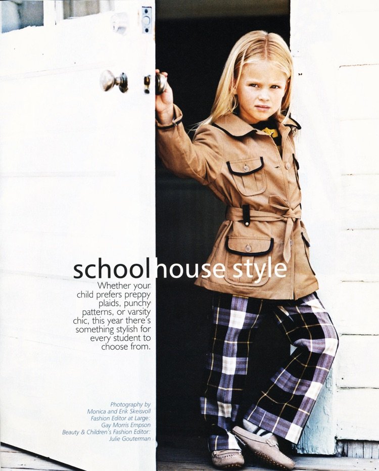A young girl leaning against a doorway, wearing a brown schoolhouse-style jacket and plaid pants.